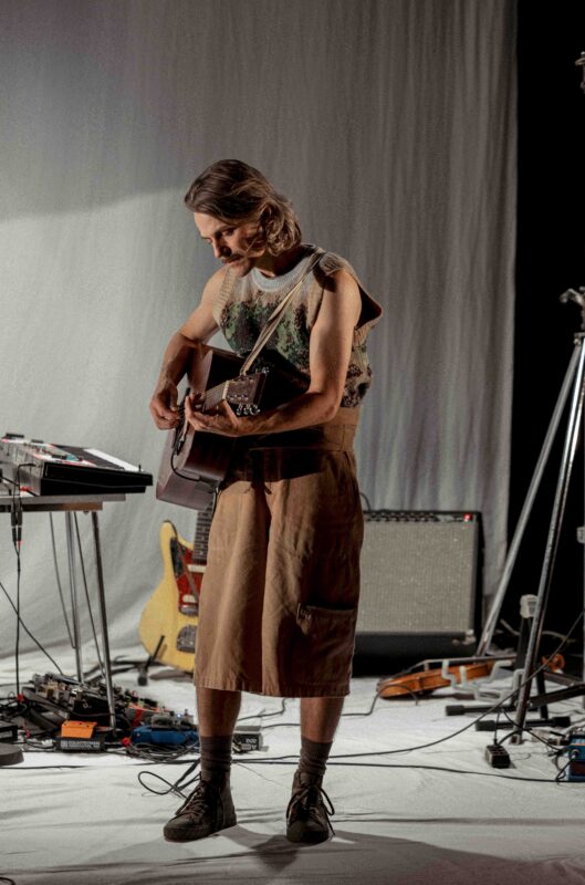 En person spiller på en akustisk gitar. Bak personen er det kabler, pedaler til gitarer og andre instrumenter.