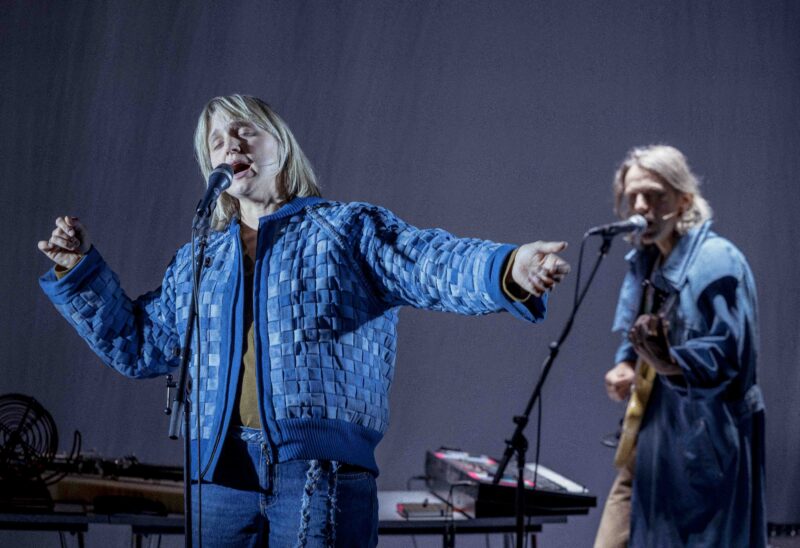 En person synger inn i en mikrofon. Bak står en annen person og spiller gitar og synger inn i en mikrofon