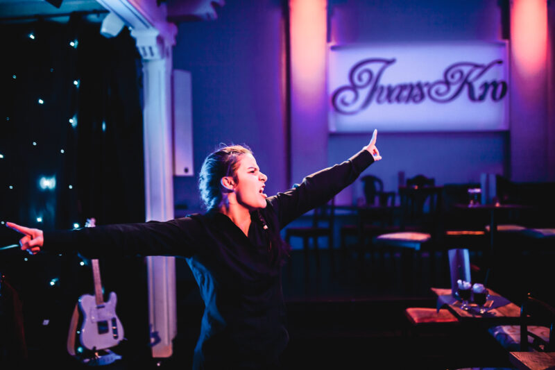 Person står skrått med armene ute til hver sin side. I bakgrunnen står det et skilt med 