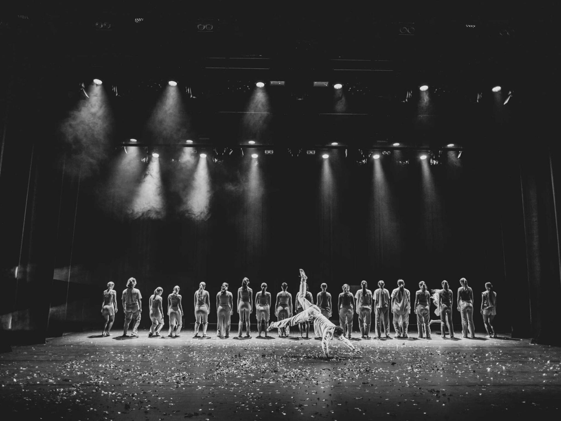 Danseensemble i rekke på scenen med ryggen til, 19 stykker. En i forgrunnen