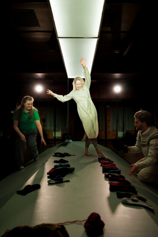 En kvinne danser på en liten rektangulær plattform med votter på. En annen kvinne står på den ene siden av plattformen og en mann sitter ser opp på kvinnen på plattformen fra den andre siden.