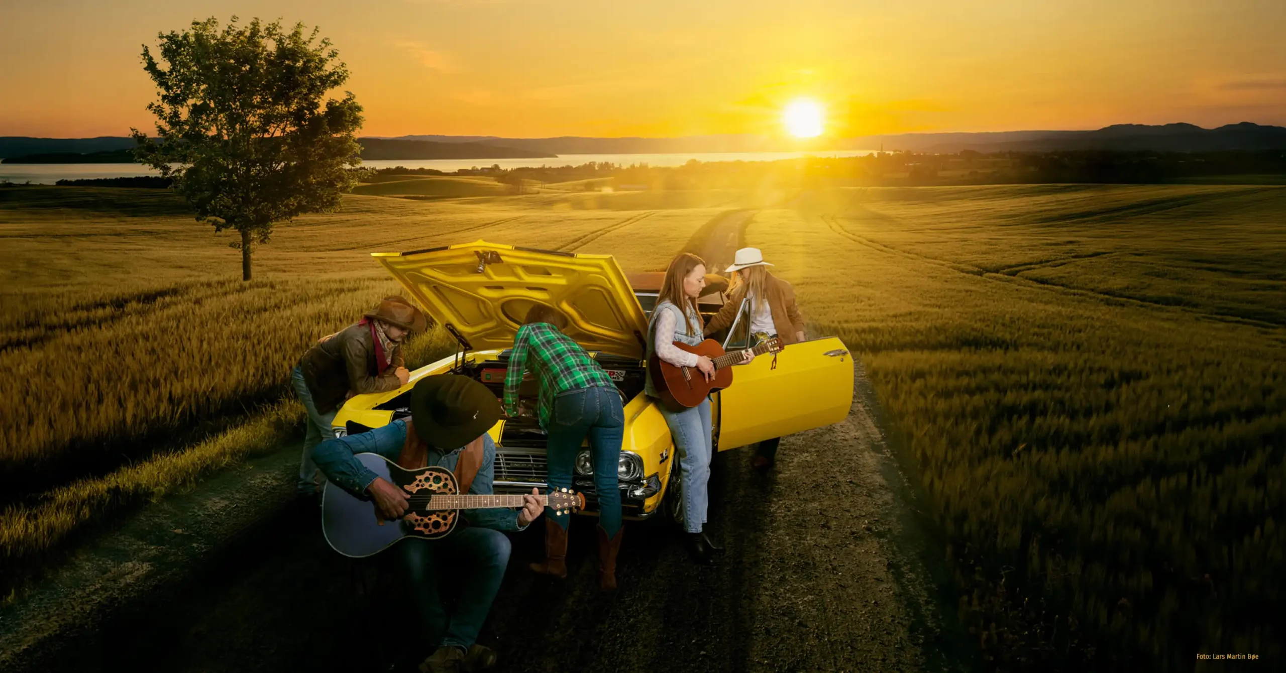 Fem personer rundt en gul bil i solnedgang. Panseret er åpent, to titter ned i panseret, to ser andre veier og spiller gitar, en annen er på vei inn i bilen.