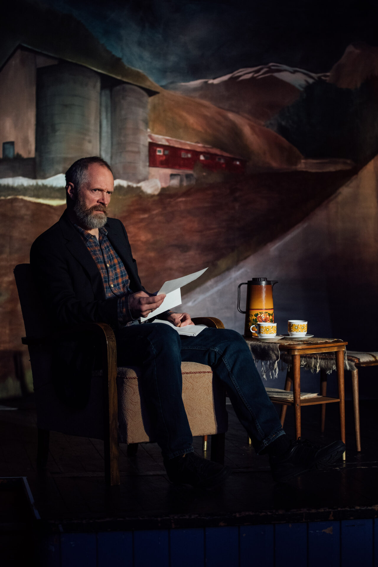 En mann med rutete skjorte og dressjakke sitter på en stol og holder et papir i den ene hånden. Ved siden av han står et lite bord med to kopper og en kaffekanne