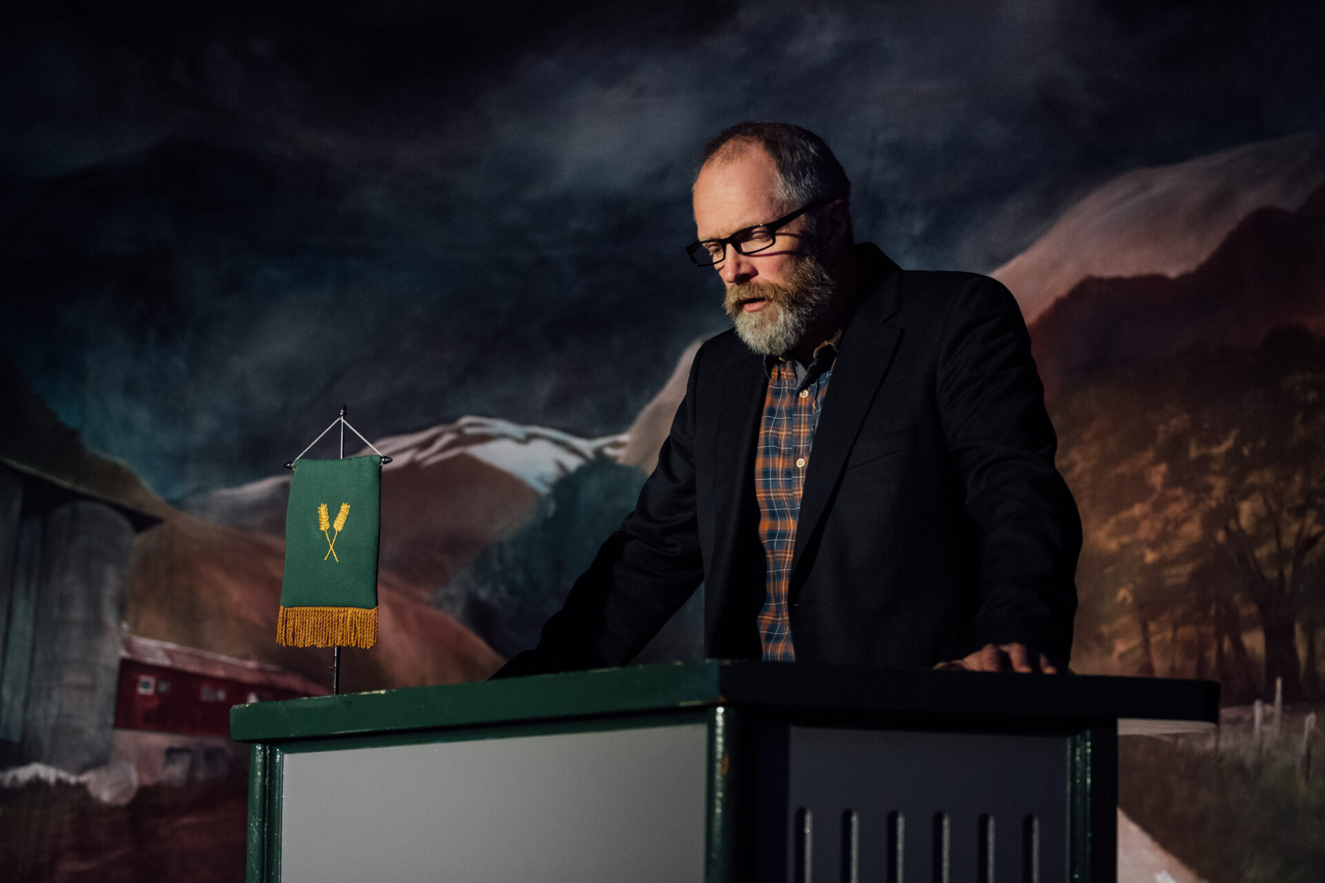 En mann med rutete skjorte og dressjakke og briller står bak en talerstol