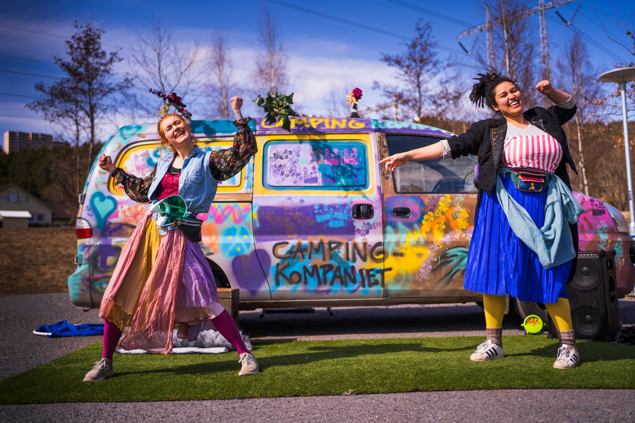 To kvinner i fargerike klær danser/jubler foran en fargerik bil.